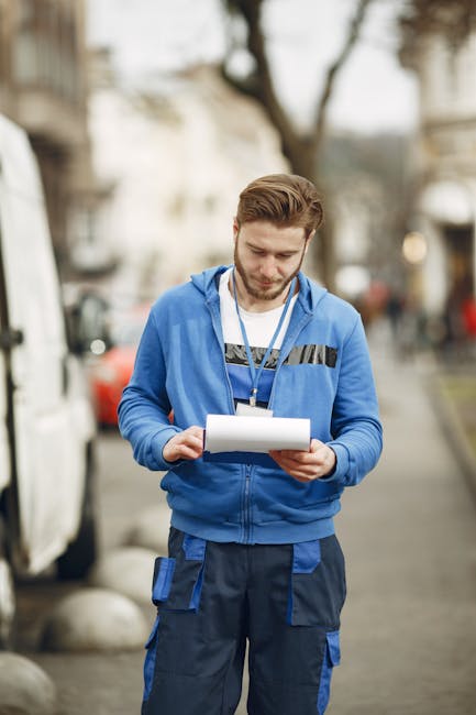 A young man with light brown hair and a beard, wearing a blue hoodie with black and grey details and blue work trousers with visible pockets, stands outdoors on a street with a blurred background of t