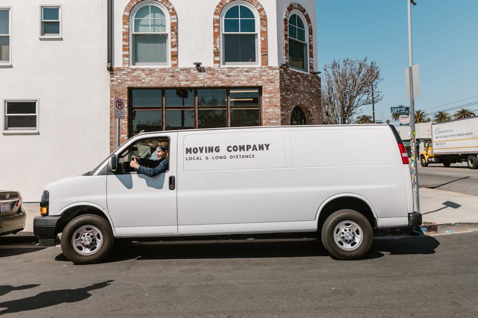 A white moving van with 'MOVING COMPANY' and 'LOCAL & LONG DISTANCE' written on the side is parked on a street in front of a brick building with large windows and an arched upper facade. The van's dri
